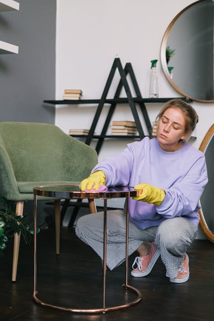 A person in a dark jacket is using a handheld vacuum cleaner to perform surface cleaning and deep cleaning on a decorative yellow and white patterned cushion, placed on a dark fabric sofa in a well-li